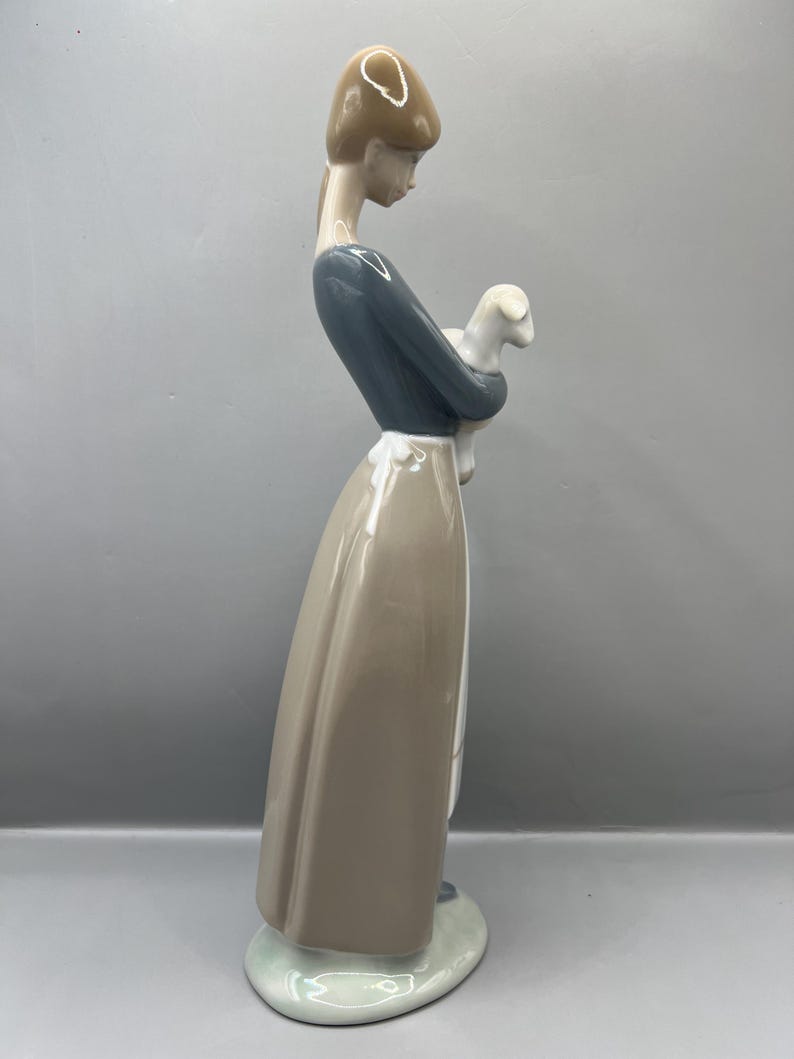 May include: A porcelain figurine of a woman in a blue dress and brown skirt holding a white lamb.