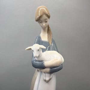 May include: A porcelain figurine of a woman in a blue and white dress holding a white lamb. The woman has brown hair and is looking down at the lamb.