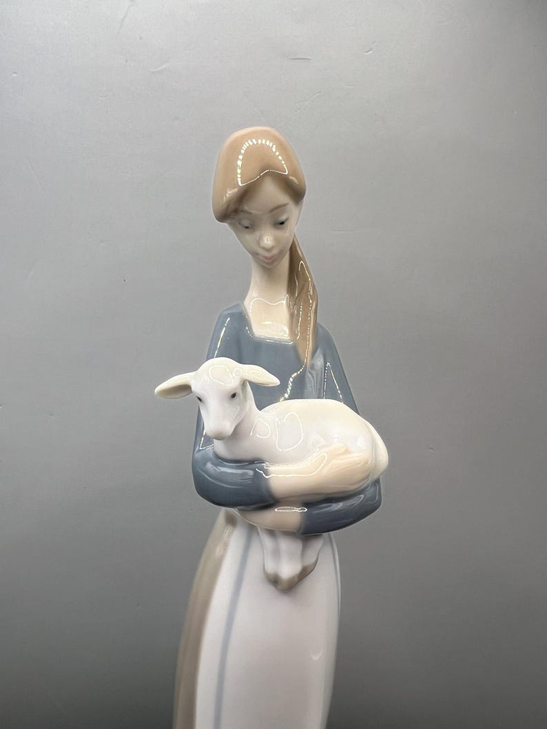 May include: A porcelain figurine of a woman in a blue and white dress holding a white lamb. The woman has brown hair and is looking down at the lamb.
