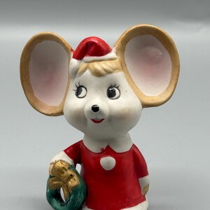 May include: A ceramic mouse figurine wearing a red Santa hat and dress, holding a green and gold wreath. The mouse has large ears, a white face, and rosy cheeks. A white collar and a white button adorn the dress.