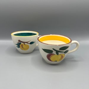 May include: Two white ceramic teacups with a yellow rim and a fruit design. One cup has a green rim and a peach design, the other has a yellow rim and an apple design.
