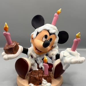 Pu&ograve; includere: Una statuina di Topolino che festeggia un compleanno. Topolino &egrave; circondato da candele rosa con fiamme gialle e torte al cioccolato. La statuina ha un design di glassa bianca e si trova su una base beige.