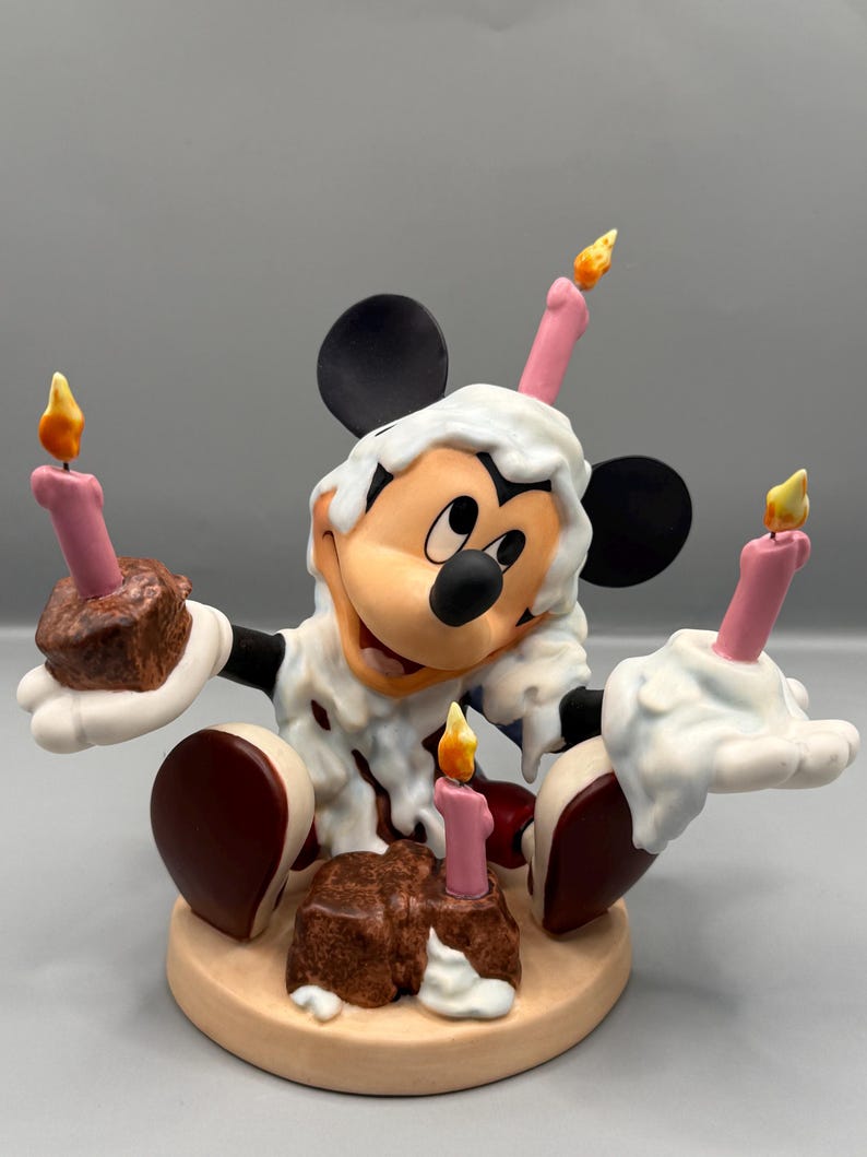 Pu&ograve; includere: Una statuina di Topolino che festeggia un compleanno. Topolino &egrave; circondato da candele rosa con fiamme gialle e torte al cioccolato. La statuina ha un design di glassa bianca e si trova su una base beige.