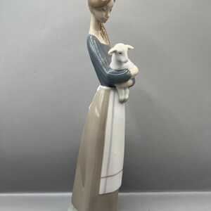 May include: A porcelain figurine of a woman in a brown dress and blue shirt holding a white lamb. The figurine is standing on a green base.