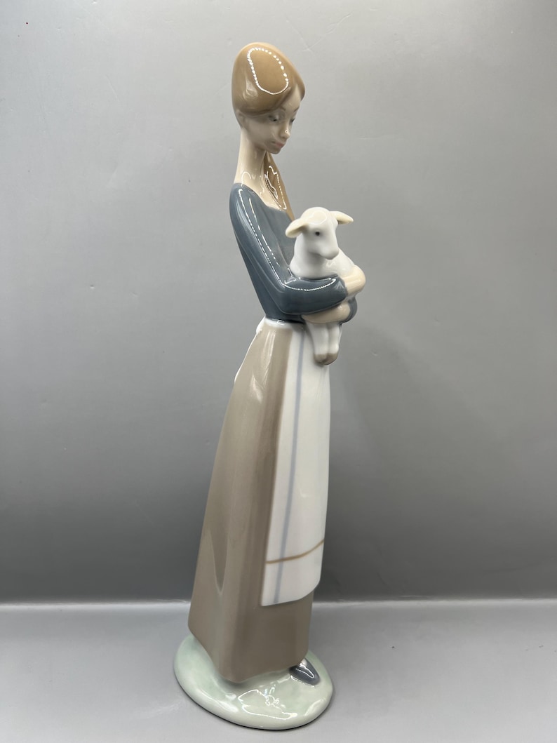 May include: A porcelain figurine of a woman in a brown dress and blue shirt holding a white lamb. The figurine is standing on a green base.
