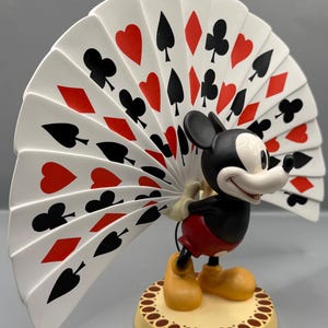 Peut inclure: Figurine de Mickey Mouse tenant un &eacute;ventail de cartes &agrave; jouer. L'&eacute;ventail affiche des c&oelig;urs rouges, des piques, des tr&egrave;fles et des carreaux noirs. Mickey est dans sa tenue classique, debout sur une base cr&egrave;me avec une bordure marron.