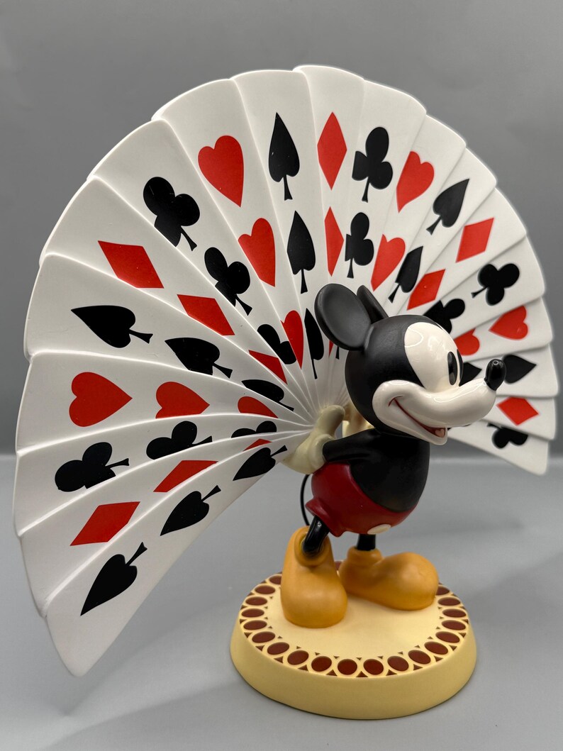 Peut inclure: Figurine de Mickey Mouse tenant un &eacute;ventail de cartes &agrave; jouer. L'&eacute;ventail affiche des c&oelig;urs rouges, des piques, des tr&egrave;fles et des carreaux noirs. Mickey est dans sa tenue classique, debout sur une base cr&egrave;me avec une bordure marron.