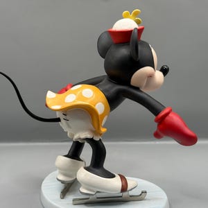 May include: A figurine of Minnie Mouse ice skating. Minnie is wearing a red hat, yellow polka-dot skirt, and red gloves. Her ice skates are white with brown accents. The figurine is on a light blue base.