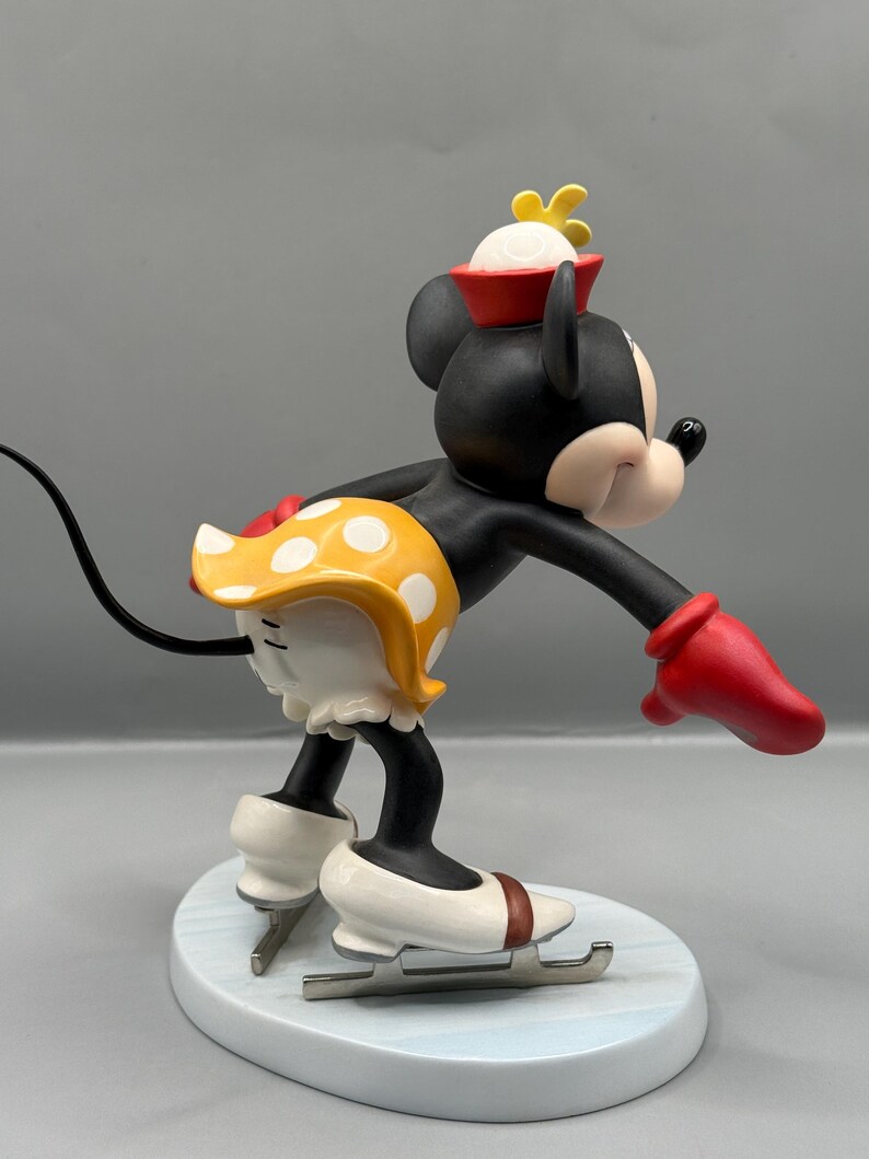 May include: A figurine of Minnie Mouse ice skating. Minnie is wearing a red hat, yellow polka-dot skirt, and red gloves. Her ice skates are white with brown accents. The figurine is on a light blue base.