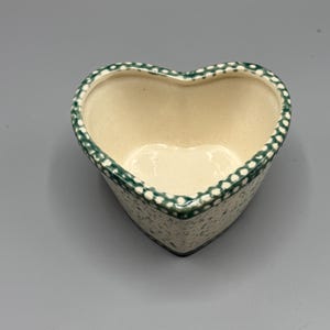 Op de afbeelding: Een hartvormige keramische schaal met een crèmekleurig interieur en een gespikkelde groen-witte buitenkant. De rand is versierd met een stippenpatroon. De lege schaal staat op een grijze ondergrond.