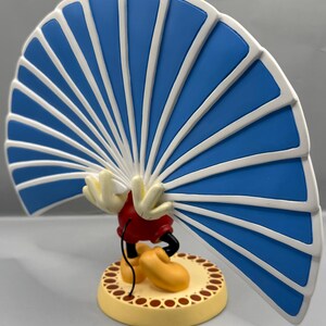 Peut inclure: Figurine d'un personnage de dessin anim&eacute; avec une grande structure en forme d'&eacute;ventail bleu et blanc. Le personnage porte un short rouge, des chaussures jaunes et des gants blancs. La base est jaune avec une bordure marron.