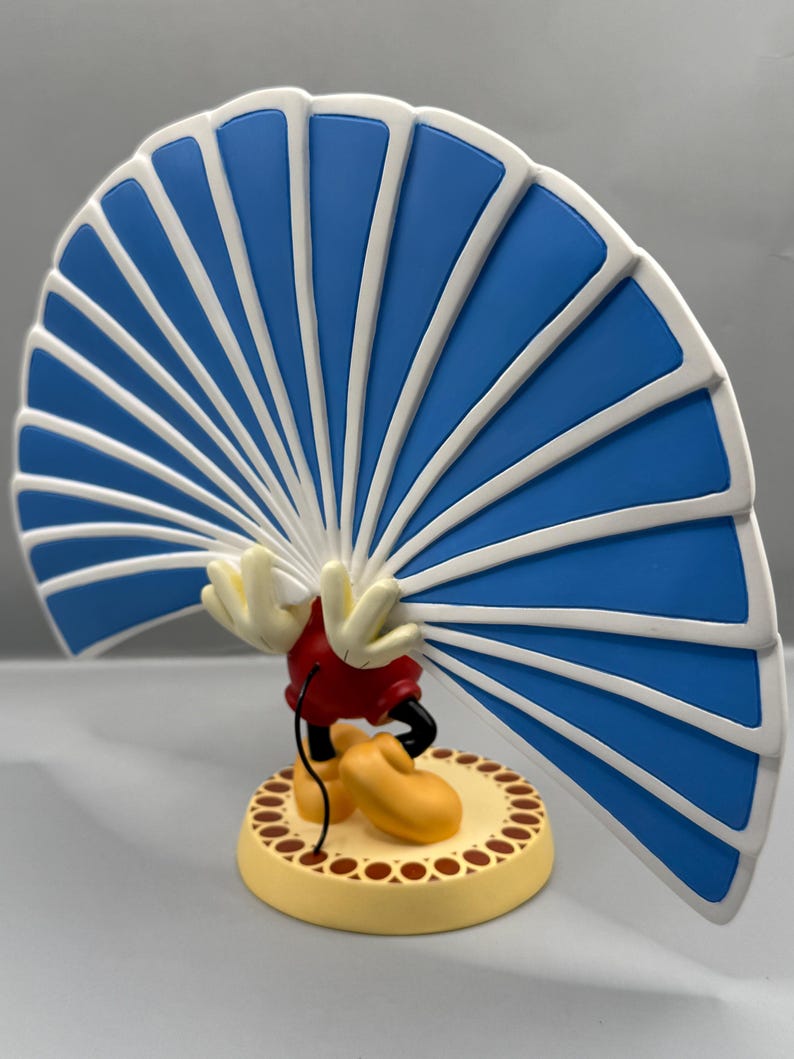 Peut inclure: Figurine d'un personnage de dessin anim&eacute; avec une grande structure en forme d'&eacute;ventail bleu et blanc. Le personnage porte un short rouge, des chaussures jaunes et des gants blancs. La base est jaune avec une bordure marron.
