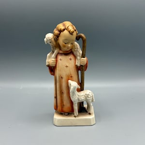 May include: A porcelain figurine of a young shepherd boy with a lamb on his shoulder and another lamb at his feet. The boy is wearing a red and gold robe with a white collar and a staff in his hand.