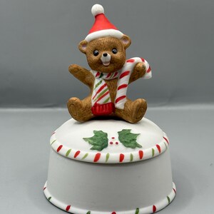 May include: A ceramic Christmas trinket box. The lid features a brown bear wearing a Santa hat and a striped scarf, holding a candy cane. The white box is decorated with red and green accents.