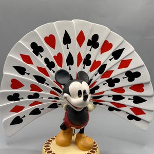 Peut inclure: Figurine de Mickey Mouse tenant un &eacute;ventail de cartes &agrave; jouer. L'&eacute;ventail est blanc avec des symboles de cartes rouges et noirs. Mickey est dans sa tenue classique, debout sur une base ronde et beige.