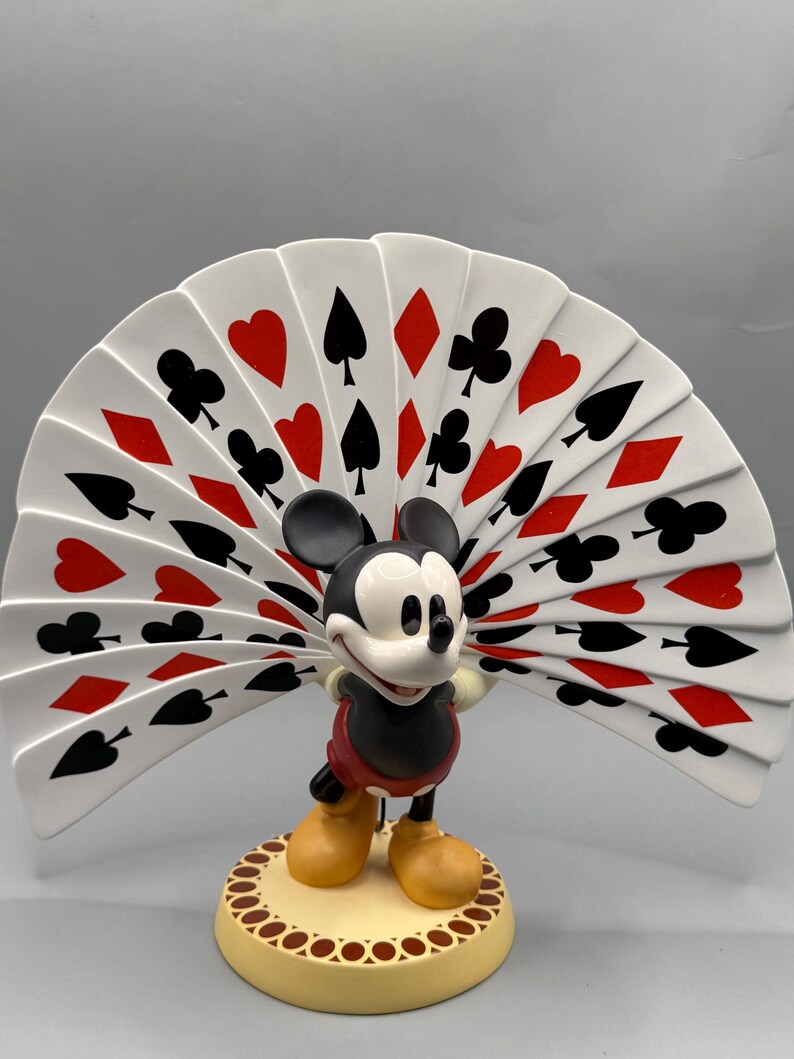 Peut inclure: Figurine de Mickey Mouse tenant un &eacute;ventail de cartes &agrave; jouer. L'&eacute;ventail est blanc avec des symboles de cartes rouges et noirs. Mickey est dans sa tenue classique, debout sur une base ronde et beige.