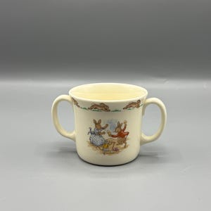 May include: Cream-colored ceramic mug with two handles, featuring a charming illustration of rabbits. The mug has a light yellow interior and a decorative border with running rabbits. A delightful piece for serving beverages.