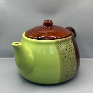 Może przedstawiać: Ceramiczny imbryk z brązową pokrywką i uchwytem, jasnozielonym korpusem oraz brązowo-zielonym, nakrapianym bokiem. Dzióbek znajduje się po lewej stronie imbryka.