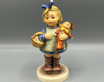 Puede incluir: Una figurita de porcelana de una niña que sostiene una muñeca. La niña lleva un vestido azul y blanco con un lazo azul en el pelo. Lleva una cesta en la mano izquierda y la muñeca en la mano derecha. La muñeca lleva un traje rojo y amarillo.
