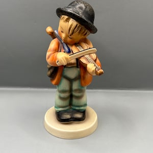 Könnte beinhalten: Eine Figur eines jungen Jungen, der einen schwarzen Hut trägt und Geige spielt. Er ist in eine braune Jacke, eine blaue Hose und schwarze Schuhe gekleidet. Die Figur ist aus Porzellan und hat einen weißen Sockel.
