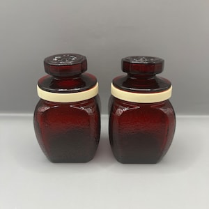 Puede incluir: Dos botellas de vidrio rojo oscuro con tapas blancas. Las botellas tienen una superficie texturizada y son cuadradas.