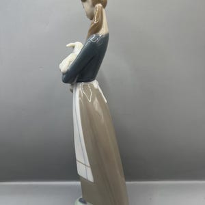 May include: A porcelain figurine of a girl in a blue and brown dress holding a white lamb. The girl has brown hair and is wearing a white apron.