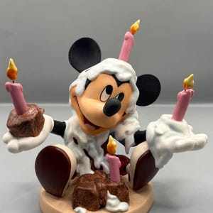 Pu&ograve; includere: Una statuina di Topolino che festeggia un compleanno. La figura &egrave; ricoperta di glassa bianca con macchie marroni, che tiene in mano due piccole torte al cioccolato con candele rosa. Due candele aggiuntive sono sulla testa. La statuina &egrave; su una base beige.