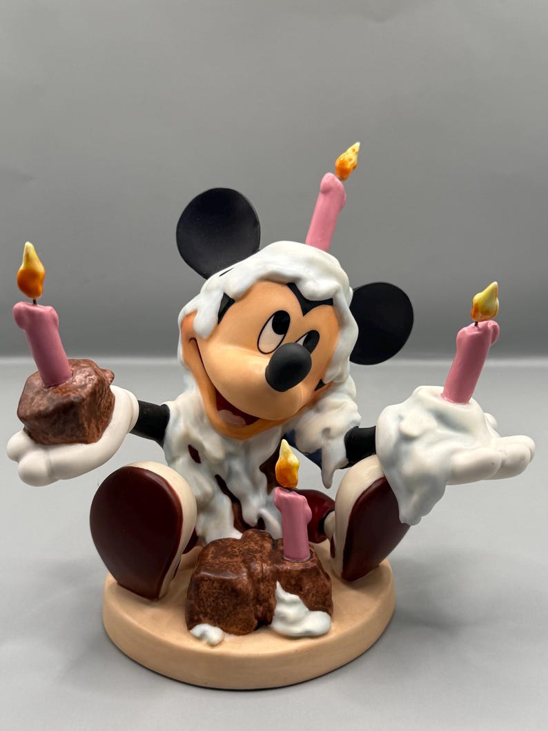Pu&ograve; includere: Una statuina di Topolino che festeggia un compleanno. La figura &egrave; ricoperta di glassa bianca con macchie marroni, che tiene in mano due piccole torte al cioccolato con candele rosa. Due candele aggiuntive sono sulla testa. La statuina &egrave; su una base beige.