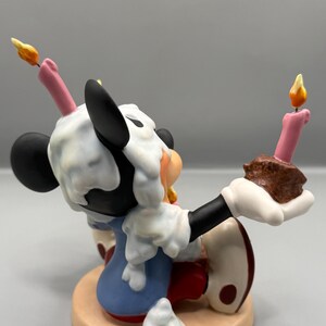 Pu&ograve; includere: Statuetta di un personaggio dei cartoni animati, con un abito blu, guanti bianchi e scarpe rosse, con orecchie nere e un viso bianco. La statuetta tiene in mano un pezzo di torta con una candela accesa e ha la glassa sulla testa. La base &egrave; beige.