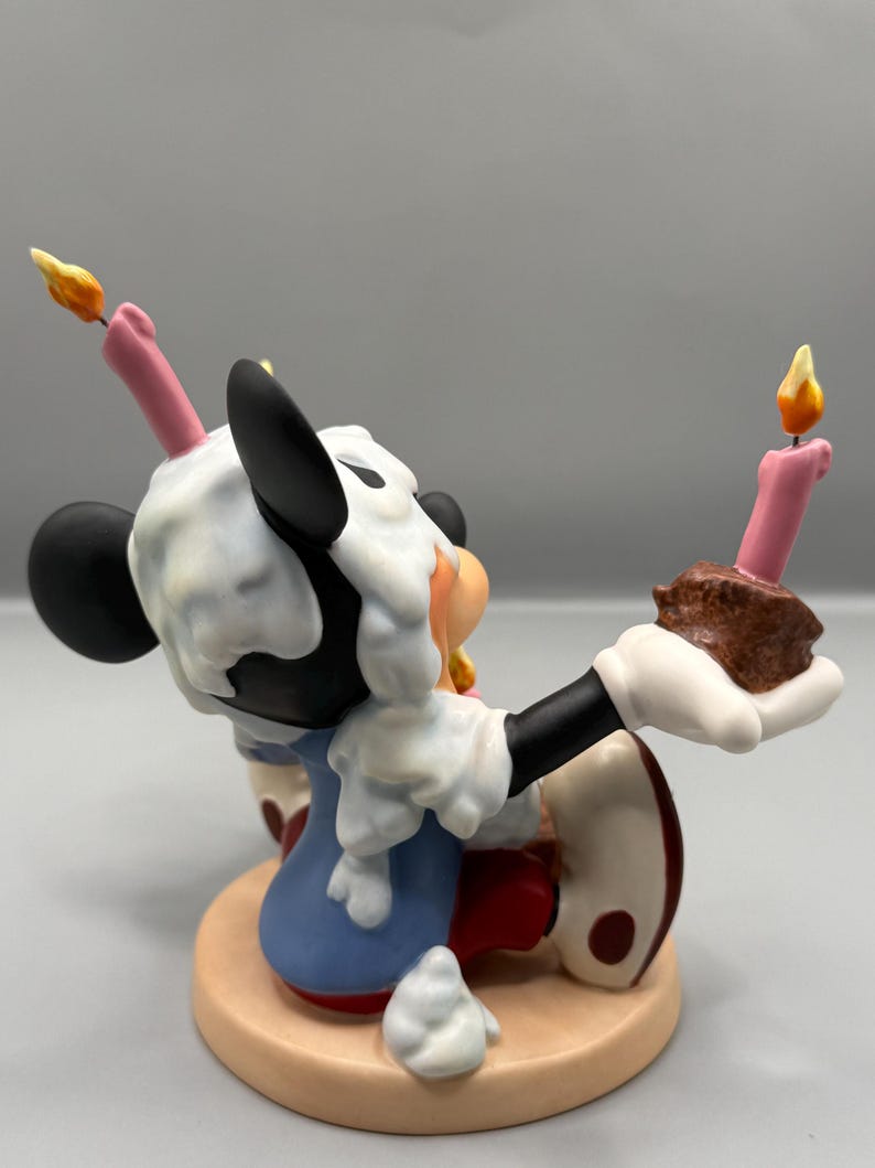 Pu&ograve; includere: Statuetta di un personaggio dei cartoni animati, con un abito blu, guanti bianchi e scarpe rosse, con orecchie nere e un viso bianco. La statuetta tiene in mano un pezzo di torta con una candela accesa e ha la glassa sulla testa. La base &egrave; beige.