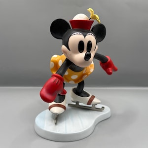 May include: A figurine of Minnie Mouse ice skating. Minnie is wearing a yellow dress with white polka dots, red gloves, and a red hat. Her ice skates are white with brown accents, and she is on a light blue base.