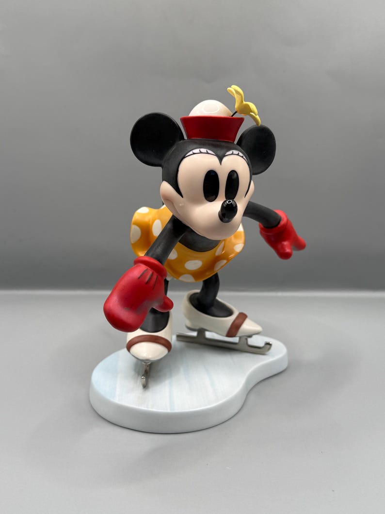 May include: A figurine of Minnie Mouse ice skating. Minnie is wearing a yellow dress with white polka dots, red gloves, and a red hat. Her ice skates are white with brown accents, and she is on a light blue base.