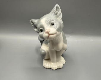 May include: A ceramic cat figurine in grey and white. The cat is seated, with a pink nose and a glossy finish. This decorative item is a charming ornament, suitable for display.