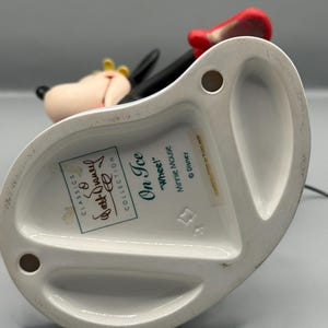 May include: A white ceramic figurine of Minnie Mouse from the "On Ice" collection. The figurine features Minnie in a skating pose, with a red bow, black dress, and yellow bow. The base has text including "Classics" and "Walt Disney Collection."