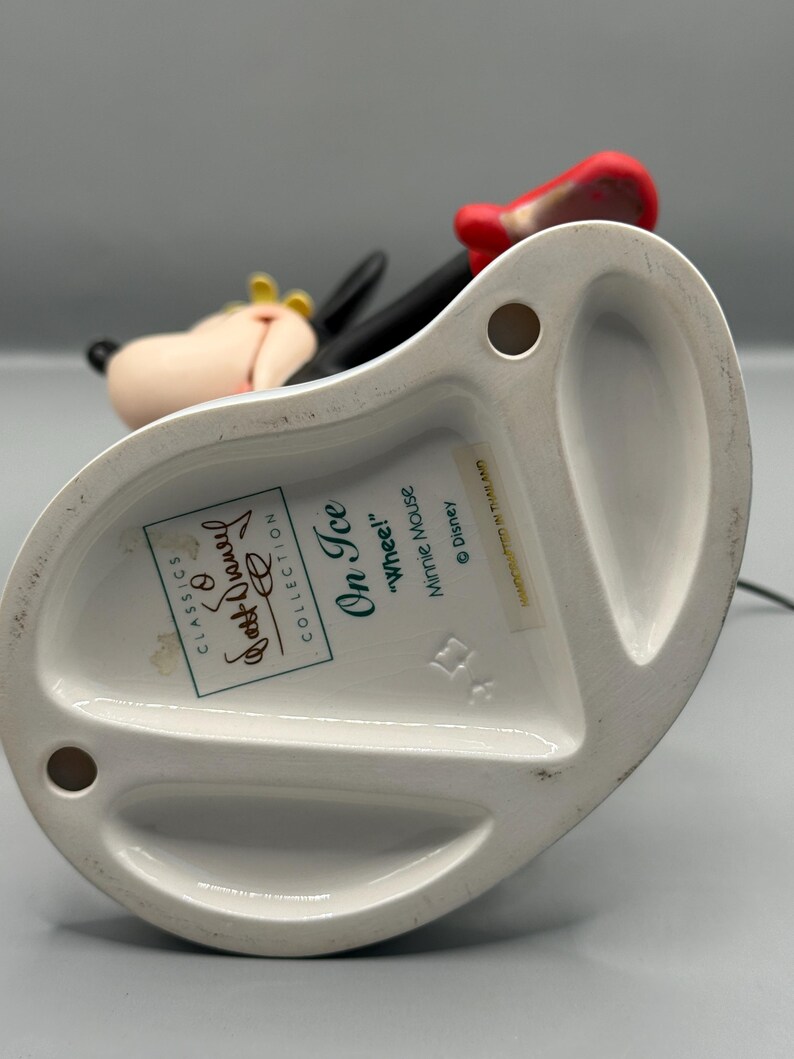 May include: A white ceramic figurine of Minnie Mouse from the "On Ice" collection. The figurine features Minnie in a skating pose, with a red bow, black dress, and yellow bow. The base has text including "Classics" and "Walt Disney Collection."