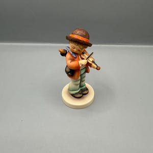 Könnte beinhalten: Figur eines jungen Geigers. Der Junge trägt einen braunen Hut mit orangefarbenem Band, eine orangefarbene Jacke und grüne Hosen. Er steht auf einem runden, cremefarbenen Sockel. Eine schwarze Tasche hängt über seiner Schulter.