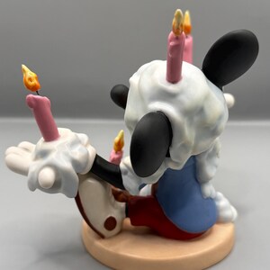Pu&ograve; includere: Una statuetta di un topo dei cartoni animati con orecchie nere, guanti bianchi e pantaloncini rossi, ricoperta di glassa bianca e candele accese. Il topo &egrave; su una base circolare color beige, con una giacca blu. Le candele hanno cera rosa e fiamme gialle.