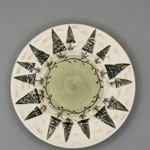 May include: A white ceramic plate with a hand-painted design. The plate features a light green center, a ring of green leaves with red berries, and a border of stylized evergreen trees in black and white. The plate has a rustic, handcrafted aesthetic.