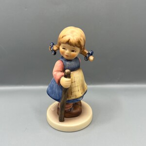 May include: A porcelain figurine of a young girl with blonde pigtails wearing a blue dress and holding a wooden stick. She is standing on a white base with the word "Hummel" written on it.