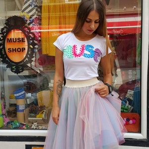 May include: A woman wearing a white t-shirt with the word "MUSE" in colourful letters and a pink tulle skirt with rainbow accents.