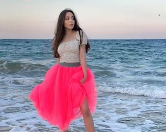 May include: A woman wearing a white top with black polka dots and a bright pink tulle skirt stands in the ocean. The skirt has a gray waistband. The background shows the ocean and a sunset.