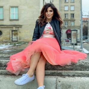 May include: A woman wearing a black leather jacket, a floral print top, and a pink tulle skirt. She is sitting on a stone ledge with her legs crossed and wearing white sneakers.