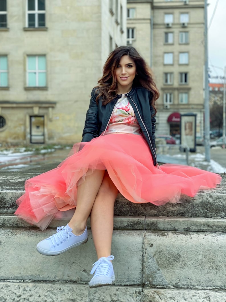 May include: A woman wearing a black leather jacket, a floral print top, and a pink tulle skirt. She is sitting on a stone ledge with her legs crossed and wearing white sneakers.