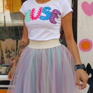 May include: A white t-shirt with the word "MUSE" printed in colourful, polka-dotted letters. The shirt is paired with a multi-coloured tulle skirt.