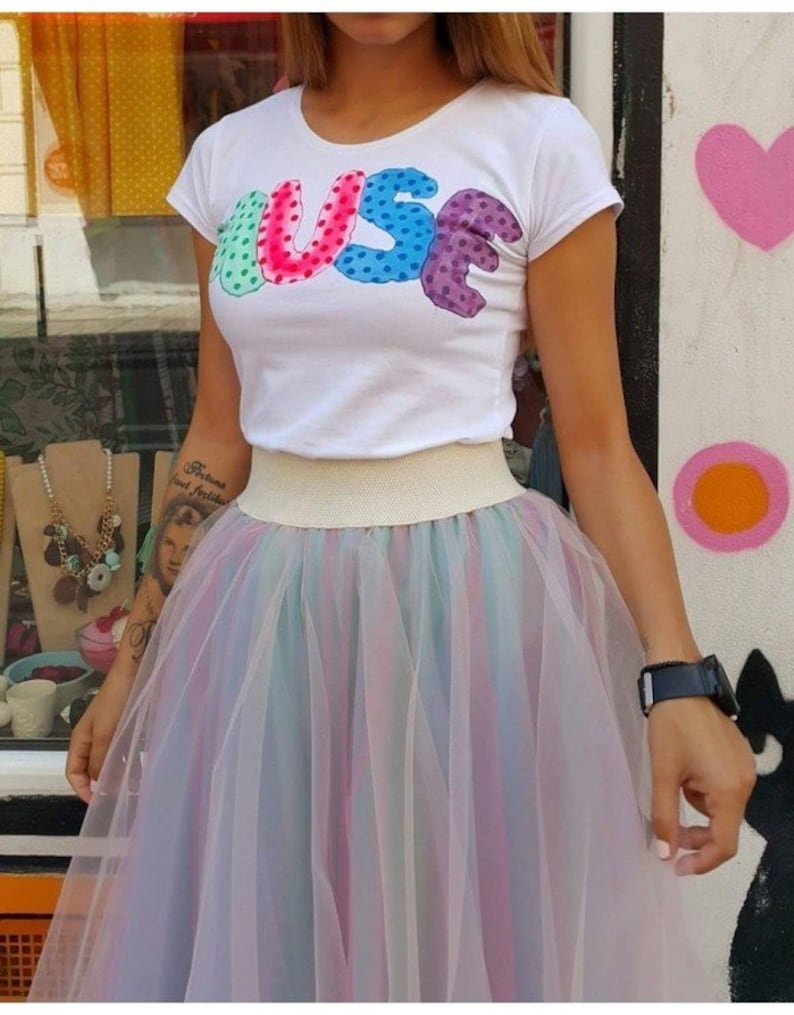 May include: A white t-shirt with the word "MUSE" printed in colourful, polka-dotted letters. The shirt is paired with a multi-coloured tulle skirt.