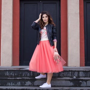 May include: A woman wearing a black leather jacket, a floral print top, and a coral tulle skirt. She is standing on a set of stairs in front of a building with black doors.