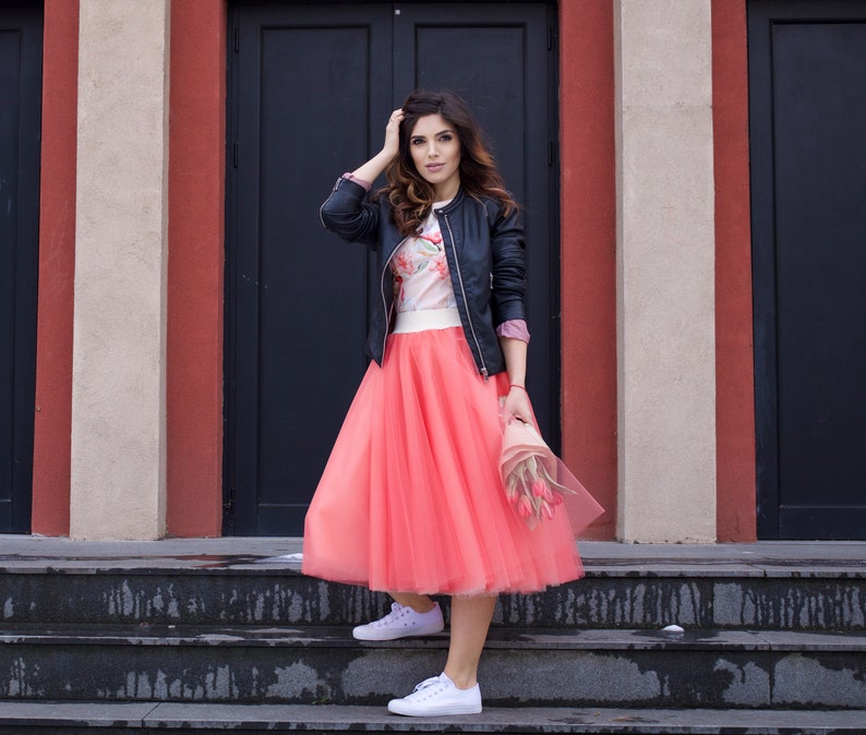 May include: A woman wearing a black leather jacket, a floral print top, and a coral tulle skirt. She is standing on a set of stairs in front of a building with black doors.