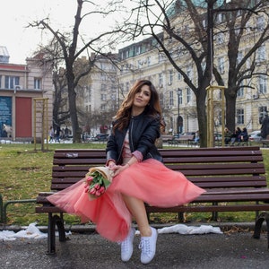 May include: A woman wearing a black leather jacket and a pink tulle skirt sits on a wooden bench holding a bouquet of pink flowers.