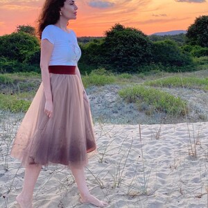 以下が含まれることがあります： 白いTシャツと茶色のチュールスカートを着た女性が、緑の草と木を背景に砂浜を歩いている。空は鮮やかなオレンジとピンクである。