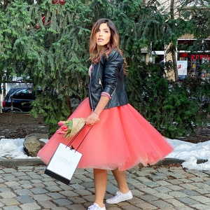 May include: A woman wearing a black leather jacket, a coral tulle skirt, and white sneakers. She is holding a bouquet of pink tulips and a white shopping bag with the word "VERNE" printed on it.